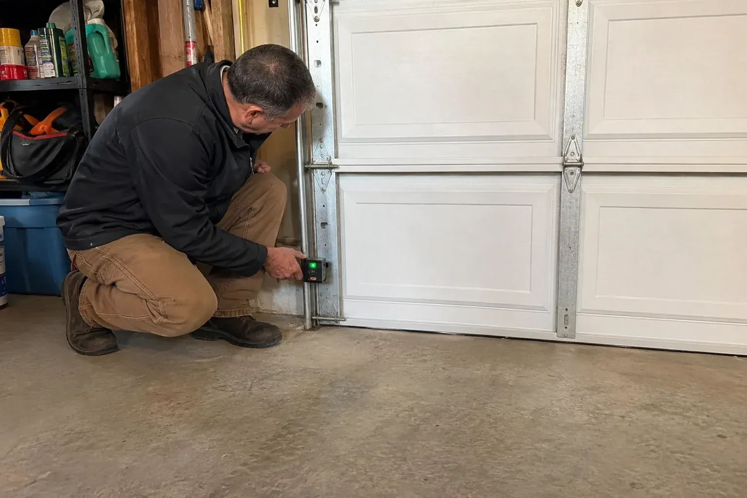 Technician kneeling beside a white paneled garage door, holding a small black box with a green light against the vertical track. — Photo-Eye Sensor Alignment or Replacement, Mississauga