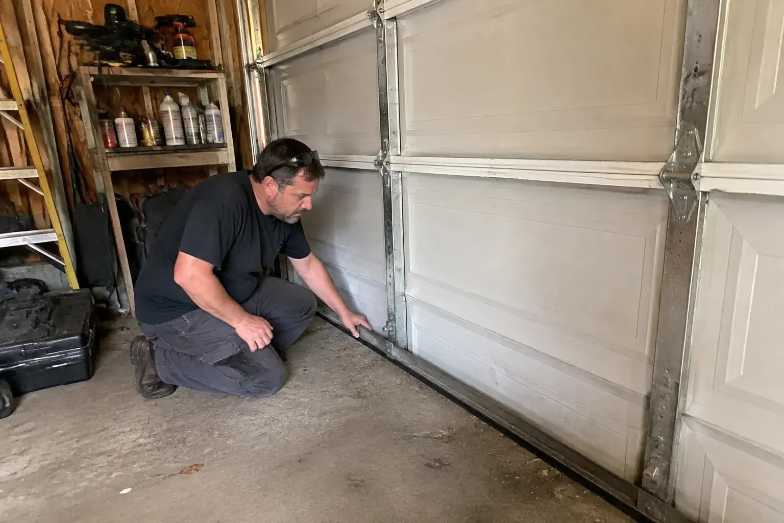 Person kneeling by a white paneled door in a garage, one hand on the black seal along its bottom edge. — Garage Door Roller Replacement, Mississauga